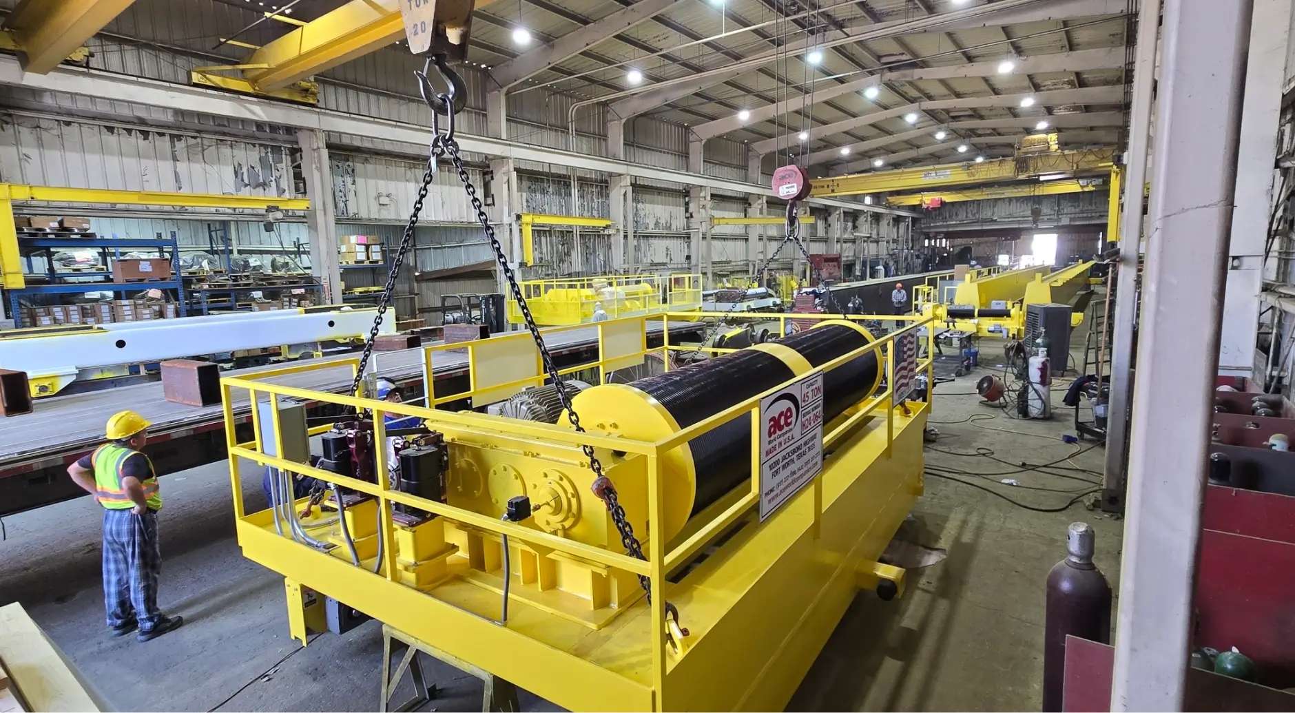 Large yellow industrial machine being lifted by an overhead crane inside a manufacturing facility.